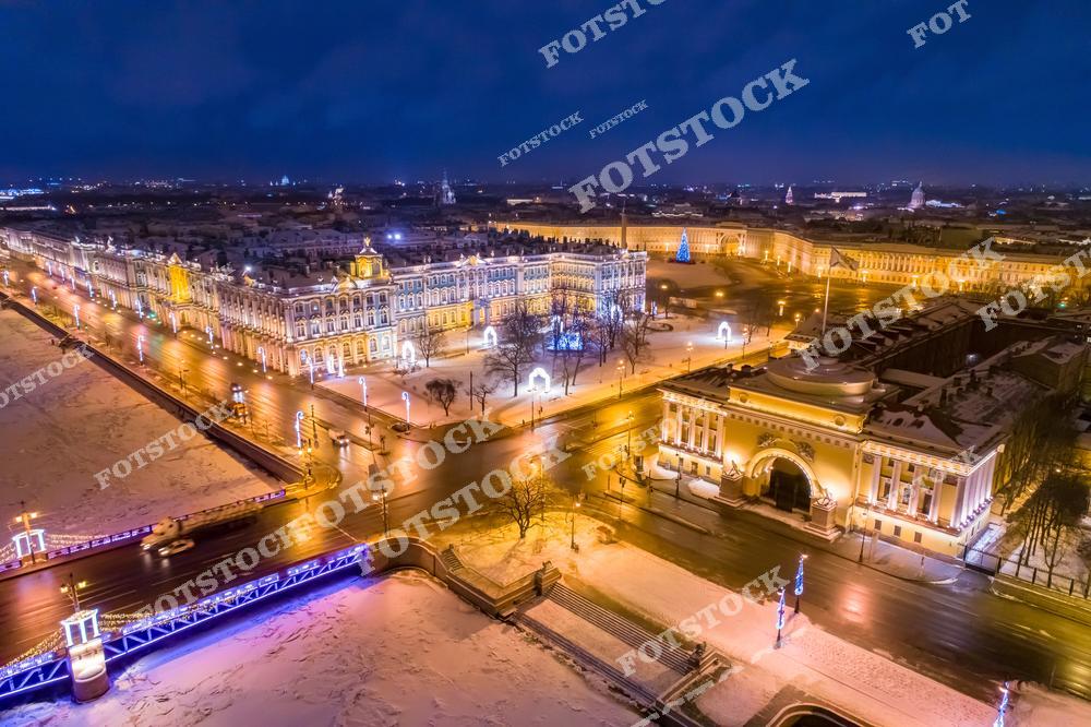 На фотографии изображена Дворцовая площадь с Зимним дворцом, частью комплекса Эрмитажа, освещенная праздничными огнями. Заснеженные улицы и елка на площади создают атмосферу зимнего праздника. На переднем плане виден Дворцовый мост с гирляндами, а вдали угадываются другие достопримечательности Санкт-Петербурга. Снимок передает всю красоту ночного Петербурга в новогодние дни.